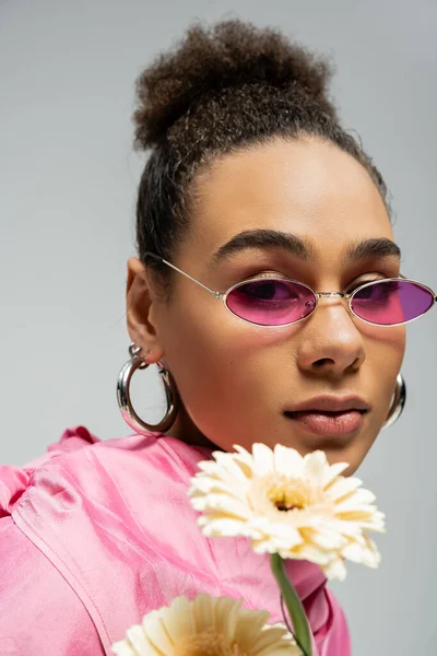 Atractiva modelo afroamericana de moda en traje rosa y gafas de sol posando con flores en gris - foto de stock