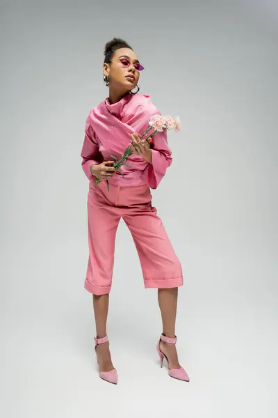 Выразительная африканская модель в розовом наряде и на каблуках, позирующая с цветами, полная длина — стоковое фото