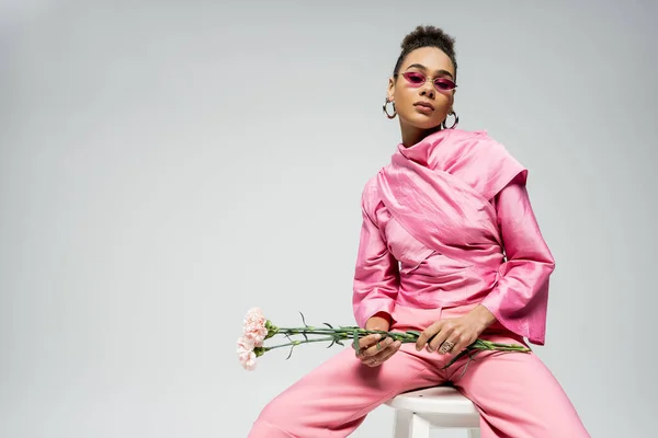 Modelo de moda americana africana em traje rosa e óculos de sol segurando flores e sentado na cadeira — Fotografia de Stock