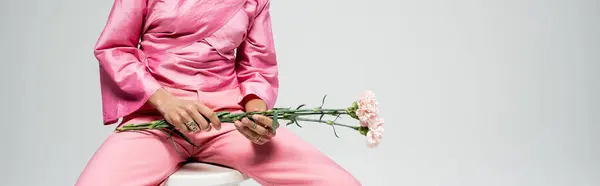 Cropped fashion model in pink attire holding flowers and sitting on chair, grey backdrop banner — Stock Photo
