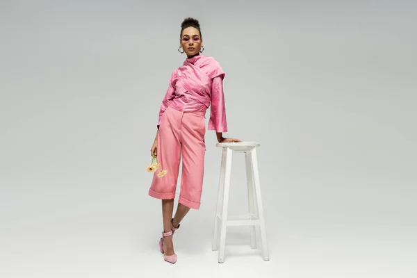 Modelo de moda americana africana em traje rosa e óculos de sol posando com flores perto de fezes em cinza — Fotografia de Stock