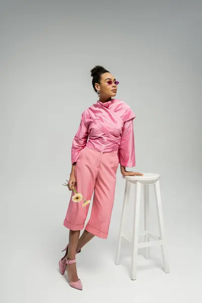 African american young woman in pink attire and sunglasses posing with flowers near stool on grey — Stock Photo