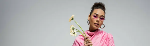 African american young woman in pink attire and sunglasses posing with flowers on grey, banner — Stock Photo
