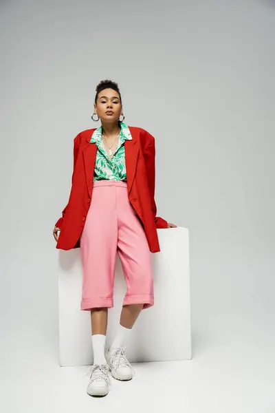 Кудрявая африканская американская модель в модном и ярком наряде позирует рядом с кубом на сером фоне — стоковое фото
