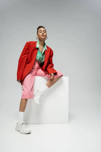 Modèle afro-américain à la mode en tenue tendance et dynamique assis sur cube sur fond gris — Photo de stock