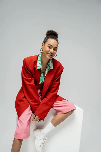Веселая африканская американская модель в модном и ярком наряде, сидящая на кубе на сером фоне — стоковое фото