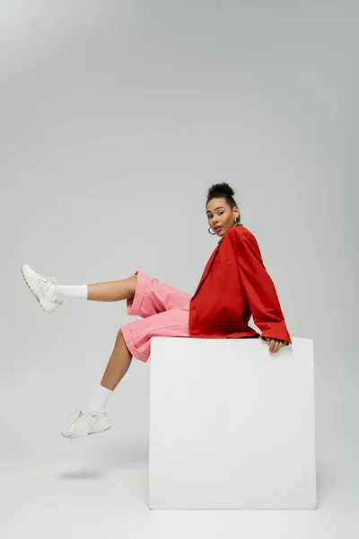 Молода афроамериканка в модному і яскравому вбранні, сидячи на кубику на сірому фоні — стокове фото