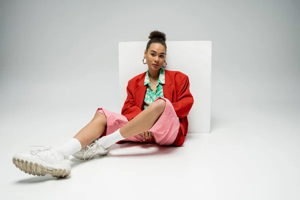Pretty african american woman in fashionable and vibrant outfit sitting near cube on grey backdrop — Stock Photo