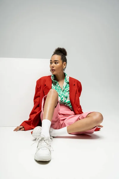 Pretty african american woman in fashionable and vibrant clothes sitting near cube on grey backdrop — Stock Photo