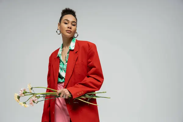 Pretty african american woman in red blazer holding flowers and looking at camera on grey backdrop — Stock Photo