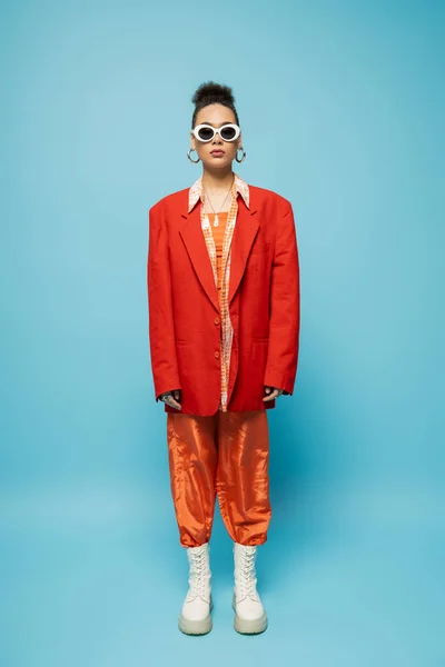 Стильна афроамериканська жінка в модних сонцезахисних окулярах і яскравому вбранні позує на синьому фоні — стокове фото