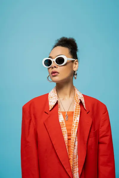 African american woman in hoop earrings, sunglasses and vibrant outfit posing on blue backdrop — Stock Photo