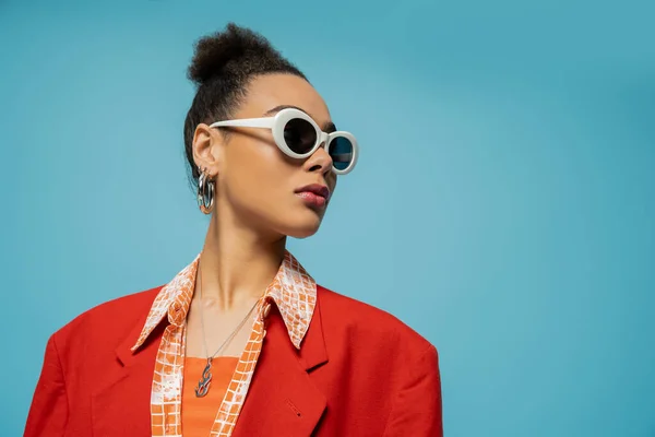Porträt einer afrikanisch-amerikanischen Frau mit Reifrohren, Sonnenbrille und lebendigem Outfit, die auf blauem Grund posiert — Stockfoto