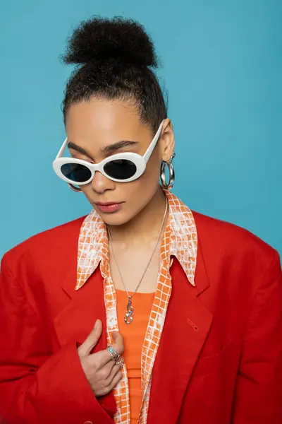 Portrait of african american woman in hoop earrings, sunglasses and vibrant outfit posing on blue — Stock Photo