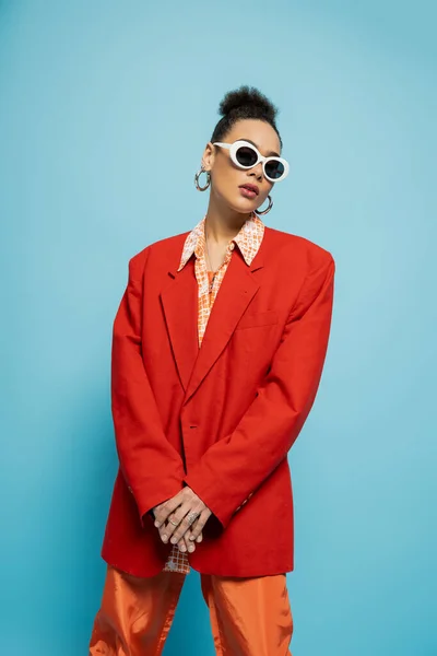 Schöne afrikanisch-amerikanische Frau in trendiger Sonnenbrille und dynamischem Outfit posiert auf blauem Hintergrund — Stockfoto