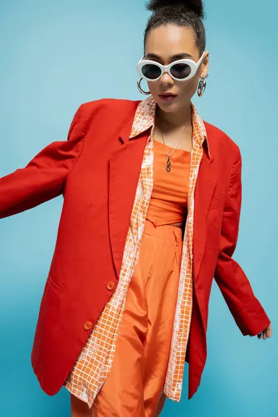 Donna afroamericana carismatica in occhiali da sole alla moda e abito vibrante in posa su sfondo blu — Foto stock