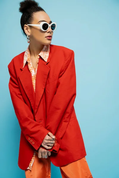 Charismatic african american model in trendy sunglasses and bold outfit posing on blue background — Stock Photo