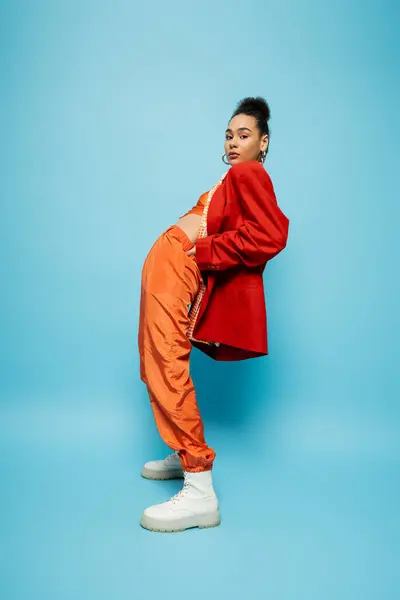 Stylish fashion model in orange suit and red blazer posing on blue backdrop and looking at camera — Stock Photo