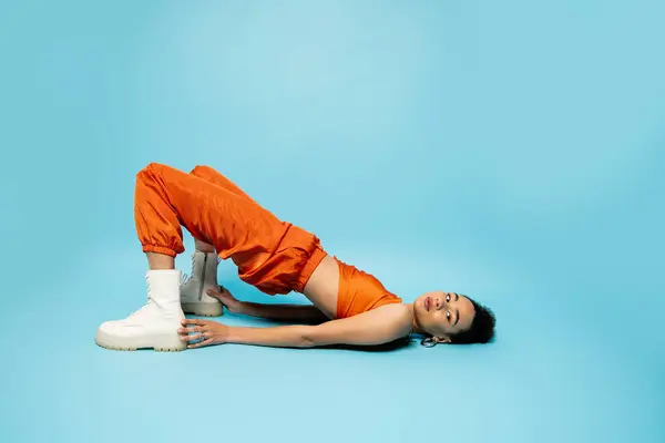 Приваблива модель моди в яскравому вуличному одязі, що лежить на підлозі, торкаючись її підборів, синій фон — стокове фото