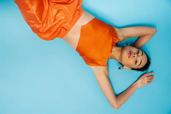 Mulher americana africana posando no chão azul vestindo roupas laranja brilhante e acessórios elegantes — Fotografia de Stock
