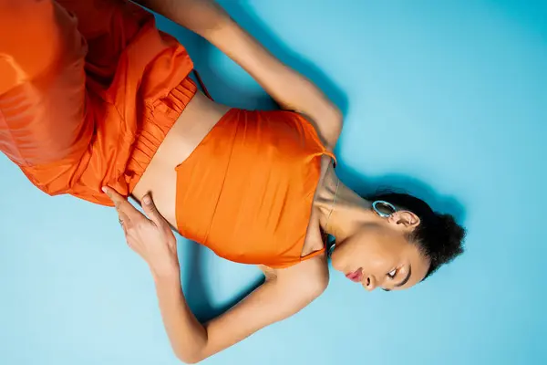 Modelo de moda afro-americano atraente posando no chão azul vestindo roupa laranja e brincos — Fotografia de Stock