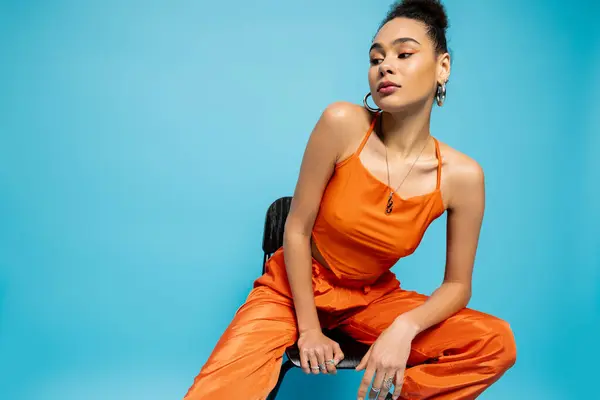 Close up attractive fashion model in orange urban suit sitting on tall chair on blue backdrop — Stock Photo