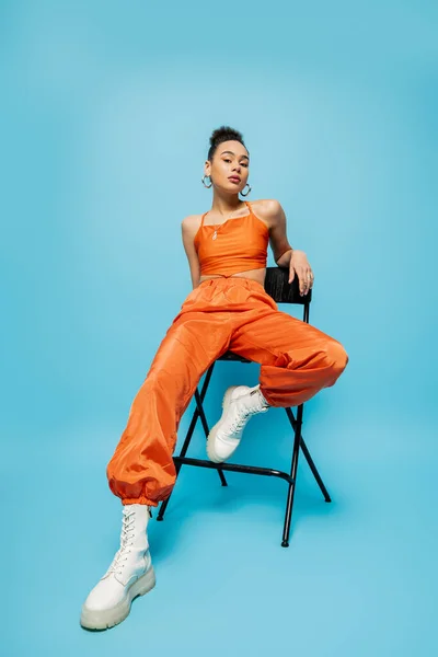Ziemlich entspanntes Mode-Modell in trendiger orangefarbener Kleidung mit Reifrohrringen auf einem hohen Stuhl sitzend — Stockfoto