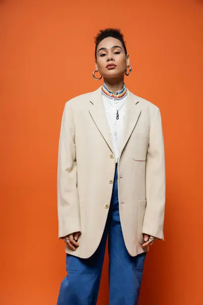 Trendy junges Mode-Model in beigem Blazer und Jeans mit Reifrohrringen posiert auf orangefarbenem Hintergrund — Stockfoto