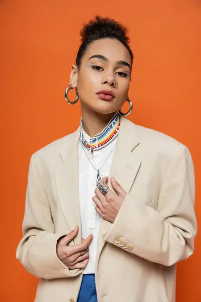 Atractiva mujer afroamericana con maquillaje vívido y accesorios en chaqueta mirando a la cámara - foto de stock