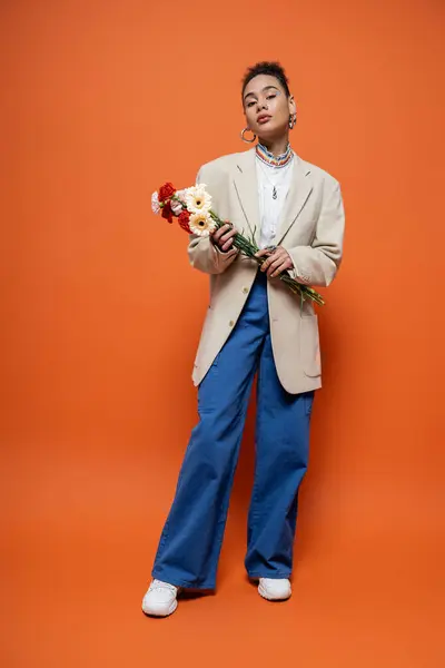 Приваблива модель моди в повсякденному міському вбранні позує на помаранчевому тлі з квітами в руках — стокове фото