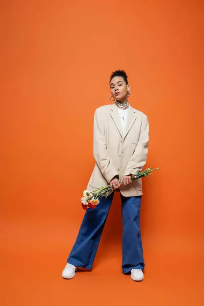 Modelo de moda elegante em calças azuis e blazer bege segurando flores em mãos em pano de fundo laranja — Fotografia de Stock