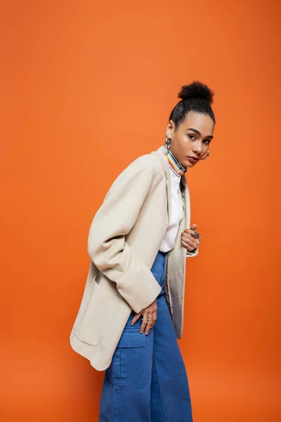 Elegant african american woman in beige blazer and blue pants looking at camera, orange backdrop — Stock Photo