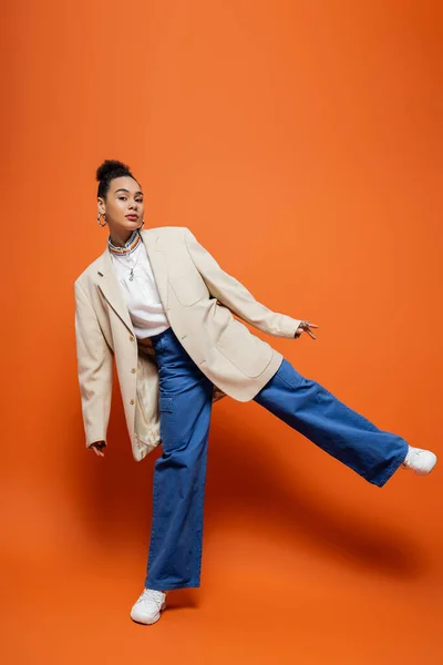 Modelo de moda elegante em blazer bege e calças azuis em pé em uma perna posando no fundo laranja — Fotografia de Stock