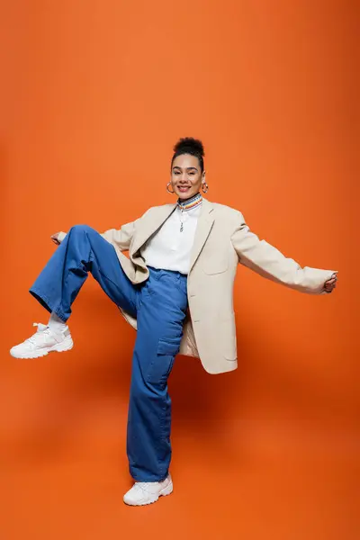 Modelo atractivo alegre en chaqueta beige y pantalones azules con accesorios posando con la rodilla levantada - foto de stock