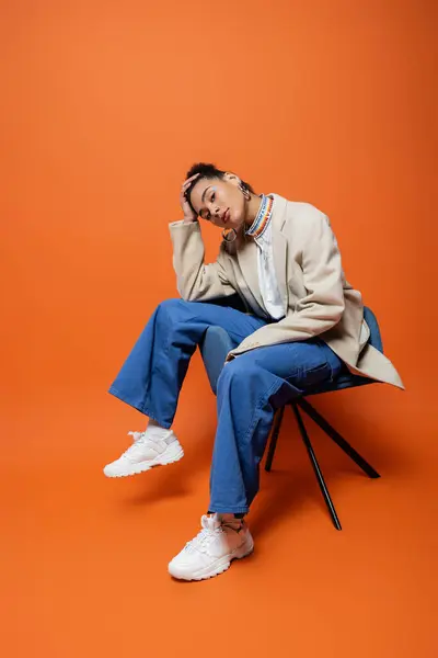 Attraktives junges Model in beigem Blazer und blauer Hose sitzt auf Stuhl und blickt in die Kamera — Stockfoto