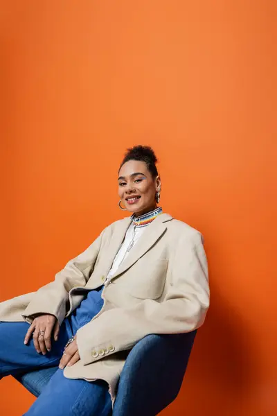 Feliz sorrindo afro-americana mulher na moda roupa sentada na cadeira e sorrindo para a câmera — Fotografia de Stock