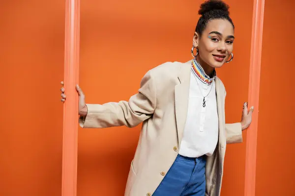 Cheerful beautiful model in stylish beige blazer and blue pants looking out of orange framework — Stock Photo