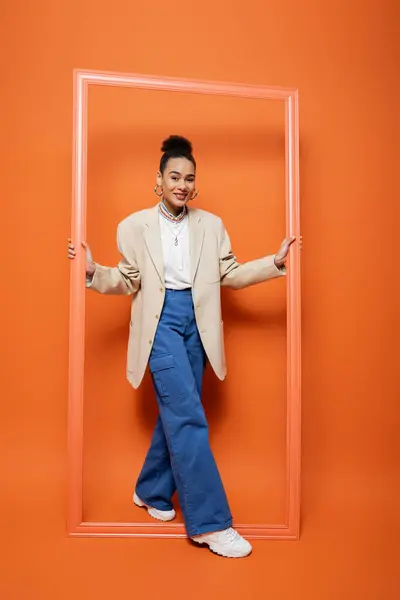 Привлекательная молодая модель в бежевом блейзере с ярким макияжем стоя и держа оранжевую рамку — стоковое фото