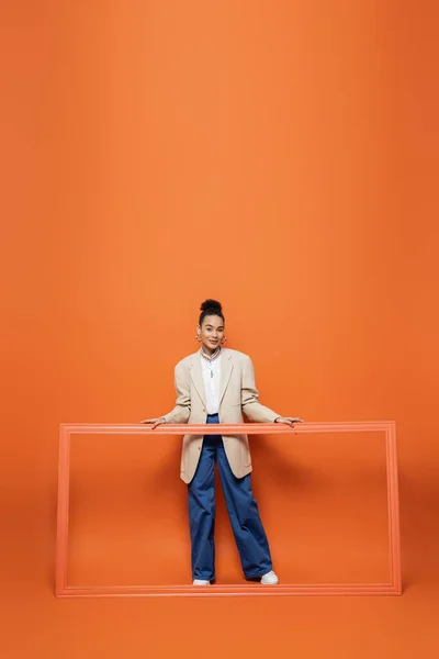 Усміхнена афро-американська модель в бежевому блістері і синіх штанах тримає рамку на помаранчевому фоні — стокове фото