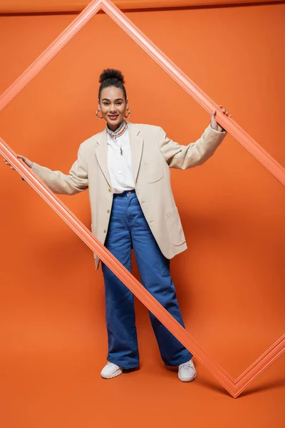 Cheerful african american fashion model in beige blazer and blue pants holding orange framework — Stock Photo