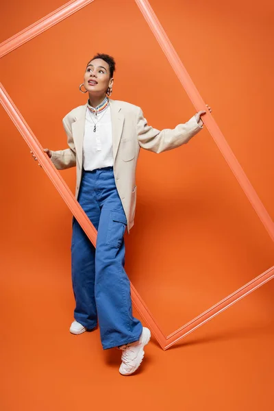 Модна жінка в бежевому блістері з сережками обруча та намистом, що позує з помаранчевою рамкою — стокове фото