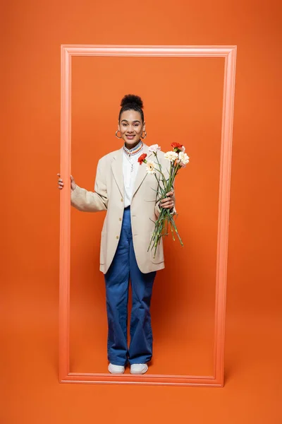 Smiling trendy african american woman in fashionable outfit holding orange frame and flower bouquet — Stock Photo