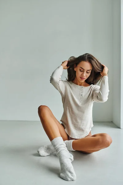 Jeune femme souriante en chaussettes et chemise à manches longues assise sur le sol à la maison et ajustant les cheveux longs — Photo de stock