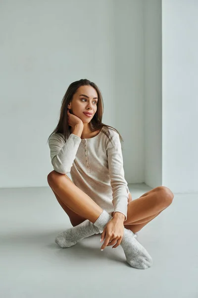 Smiling young woman in socks and long sleeve shirt sitting on floor at home and looking away — Stock Photo