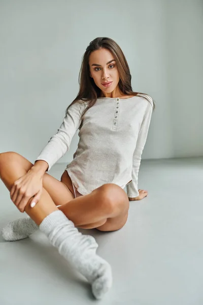 Attrayant jeune femme en chaussettes et chemise à manches longues assis sur le sol à la maison et regardant la caméra — Photo de stock