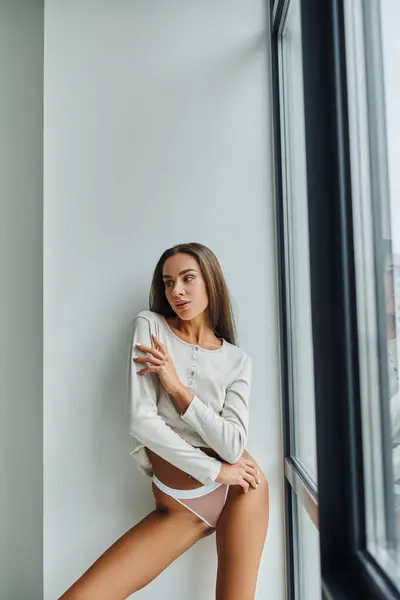 Morena mujer joven en camisa de manga larga y bragas de pie cerca de la ventana panorámica, sexy - foto de stock