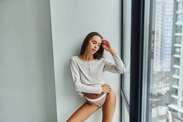 Bruna e sexy donna in camicia a maniche lunghe e mutandine in piedi vicino alla finestra panoramica — Foto stock