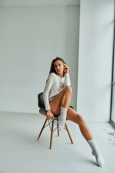 Nachdenkliche und sexy Frau in langärmeligem Hemd und Socken zu Hause auf Stuhl sitzend, Weiblichkeit — Stockfoto