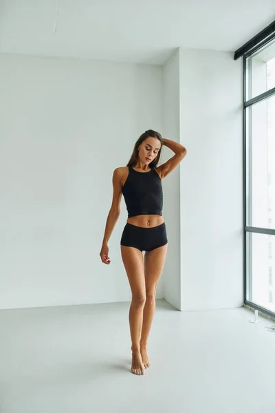 Pleine longueur de pieds nus brunette femme en sous-vêtements noirs debout près de la fenêtre panoramique à la maison — Photo de stock