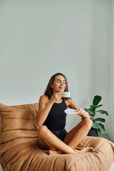 Mujer joven complacida con los ojos cerrados disfrutando de la taza de café, sentado en la silla cómoda bolsa de frijol - foto de stock